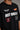Man wearing a black STAR BOY oversized t-shirt with "Hot Wheels" graphics, sitting on a black couch. Streetwear fashion.