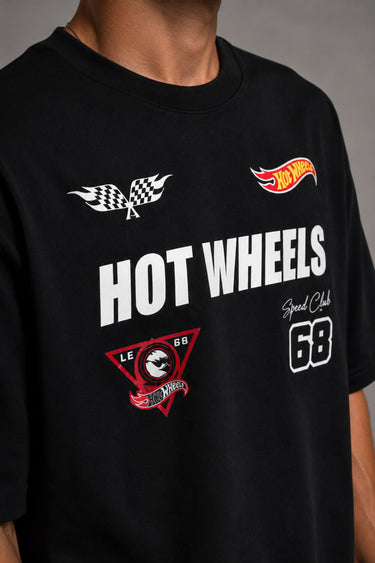 Man wearing a black STAR BOY oversized t-shirt with "Hot Wheels" graphics, sitting on a black couch. Streetwear fashion.