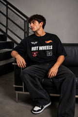Man wearing a black STAR BOY oversized t-shirt with "Hot Wheels" graphics, sitting on a black couch. Streetwear fashion.