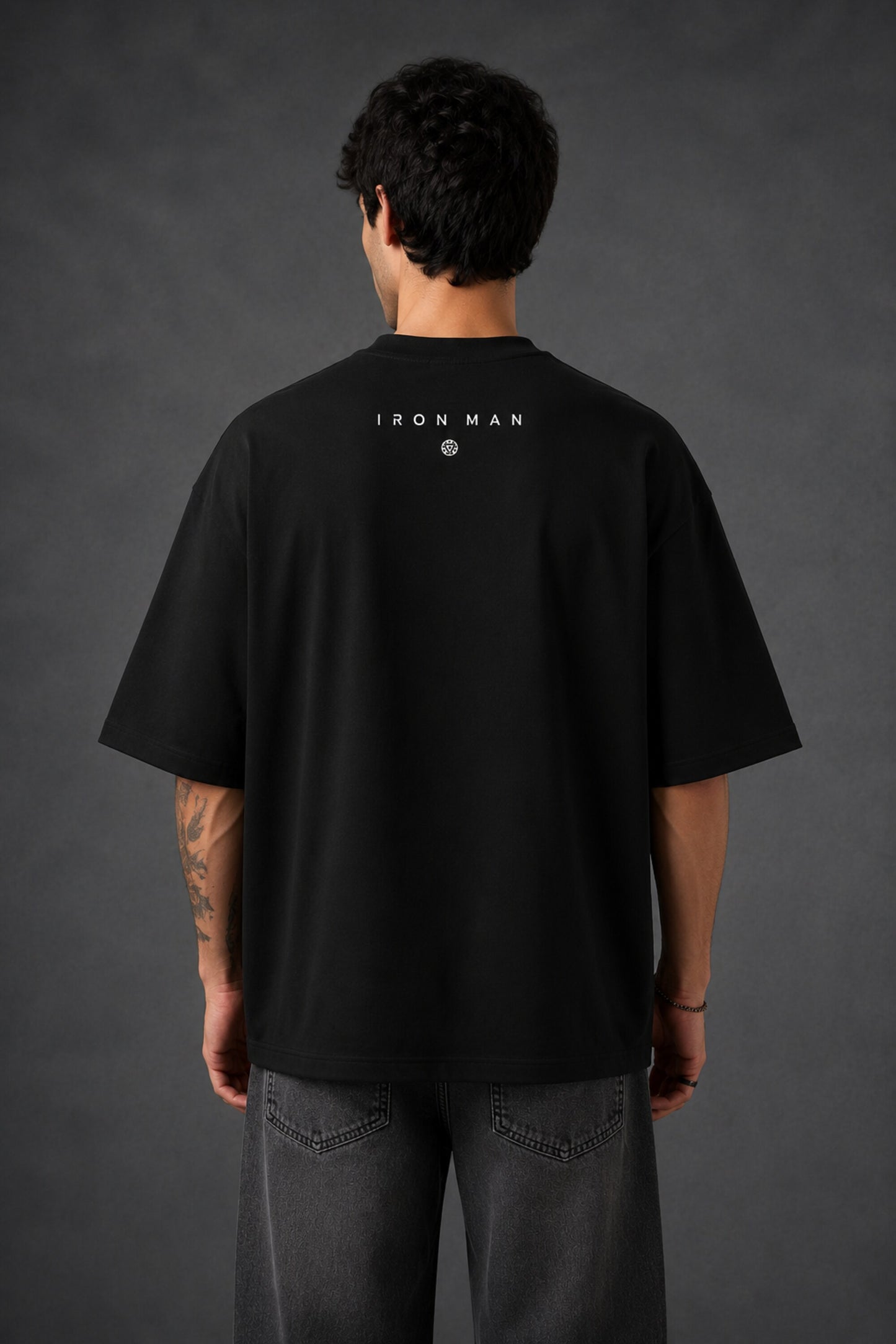 Couple wearing black Iron Man Jarvish oversized t-shirts. Man's back shows "IRON MAN" logo. Woman's front shows Iron Man arc reactor logo. Dark grey background.