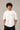 Man wearing a white 'THE KNIGHT' oversized t-shirt and black pants, sitting on concrete stairs. Features short curly hair and white sneakers. - white