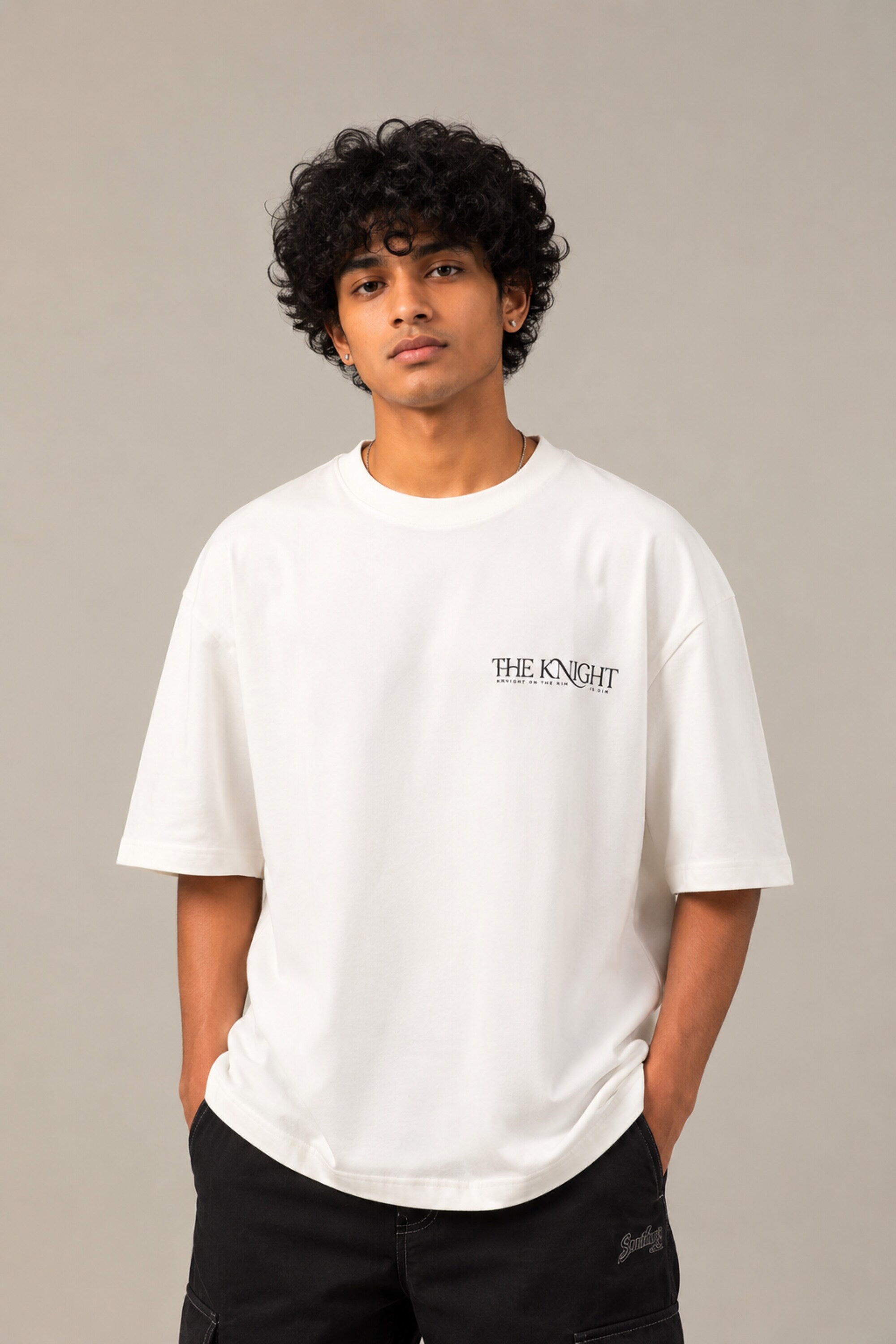 Man wearing a white 'THE KNIGHT' oversized t-shirt and black pants, sitting on concrete stairs. Features short curly hair and white sneakers. - white