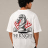Man wearing a white 'THE KNIGHT' oversized t-shirt and black pants, sitting on concrete stairs. Features short curly hair and white sneakers.