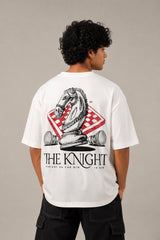 Man wearing a white 'THE KNIGHT' oversized t-shirt and black pants, sitting on concrete stairs. Features short curly hair and white sneakers.