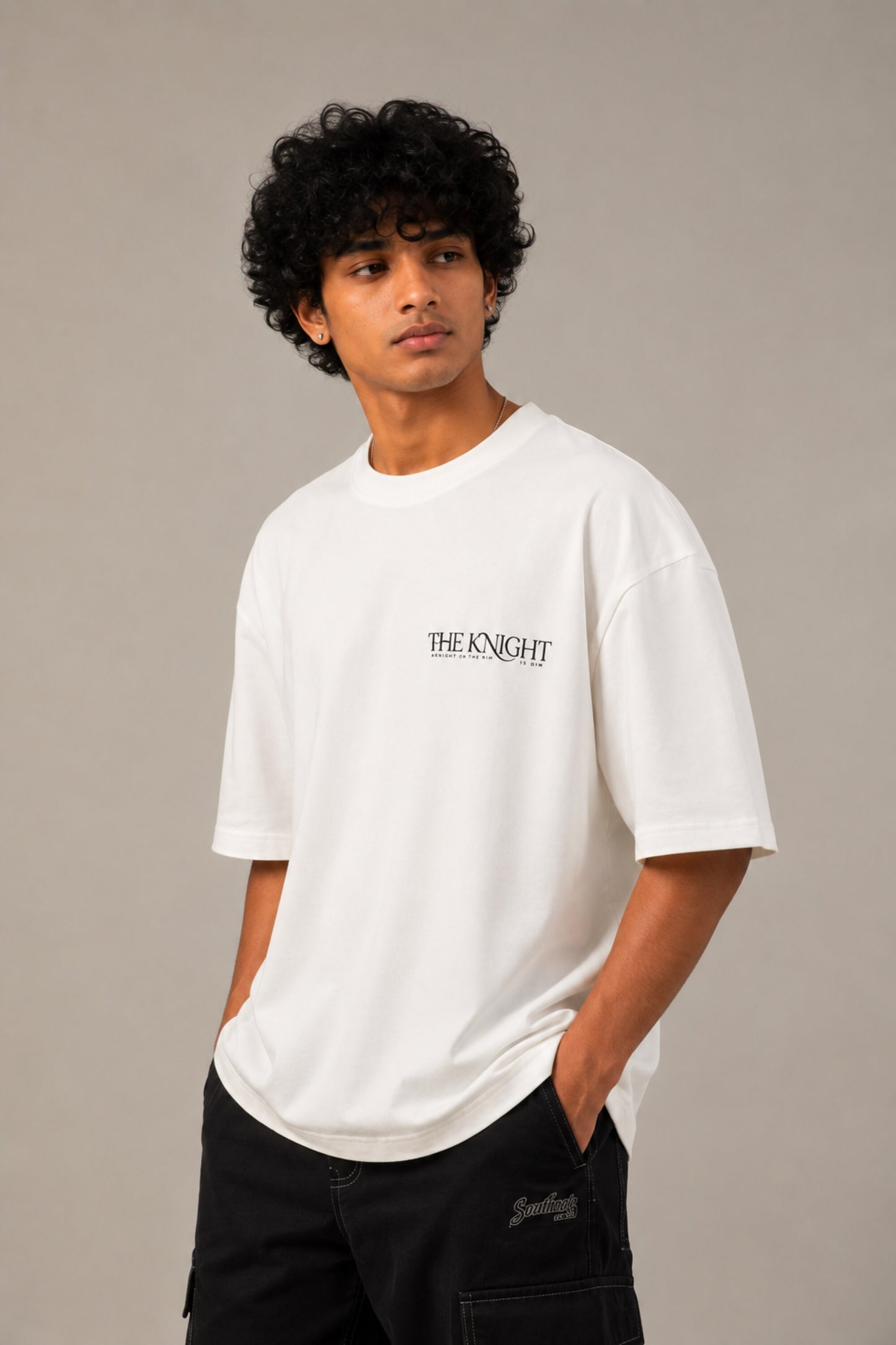 Man wearing a white 'THE KNIGHT' oversized t-shirt and black pants, sitting on concrete stairs. Features short curly hair and white sneakers.