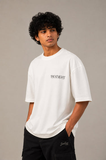 Man wearing a white 'THE KNIGHT' oversized t-shirt and black pants, sitting on concrete stairs. Features short curly hair and white sneakers.