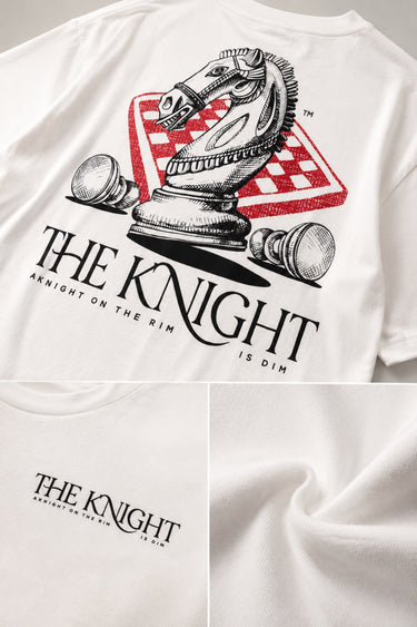 Man wearing a white 'THE KNIGHT' oversized t-shirt and black pants, sitting on concrete stairs. Features short curly hair and white sneakers.