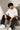 Man wearing a white 'THE KNIGHT' oversized t-shirt and black pants, sitting on concrete stairs. Features short curly hair and white sneakers.