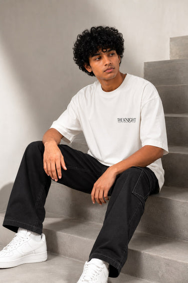 Man wearing a white 'THE KNIGHT' oversized t-shirt and black pants, sitting on concrete stairs. Features short curly hair and white sneakers.