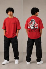 Man wearing a white 'THE KNIGHT' oversized t-shirt and black pants, sitting on concrete stairs. Features short curly hair and white sneakers. - red