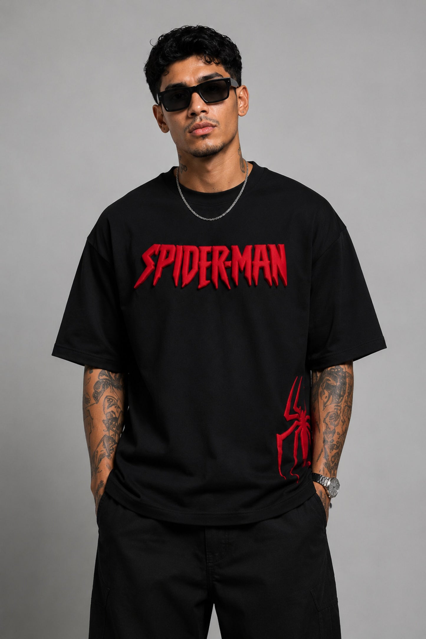 Man wearing a black oversized t-shirt with a red 'SPIDERMAN' puff print graphic and a red spider logo on the side. Model is wearing sunglasses and cargo pants.