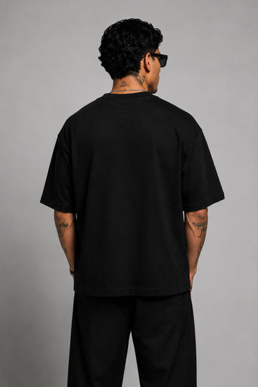 Man wearing a black oversized t-shirt with a red 'SPIDERMAN' puff print graphic and a red spider logo on the side. Model is wearing sunglasses and cargo pants.
