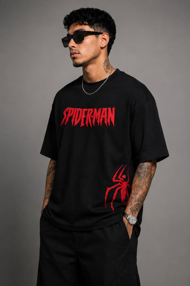 Man wearing a black oversized t-shirt with a red 'SPIDERMAN' puff print graphic and a red spider logo on the side. Model is wearing sunglasses and cargo pants.