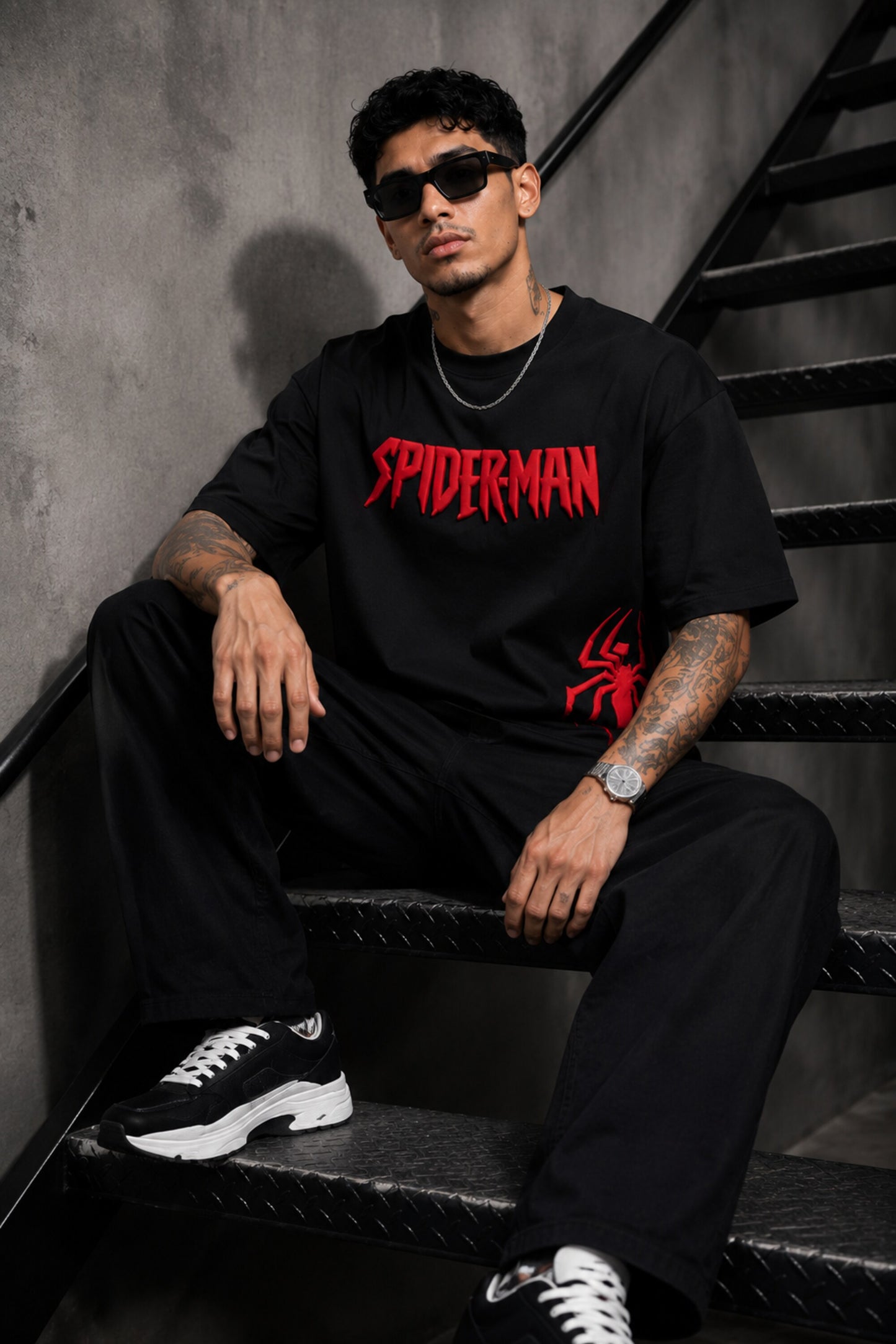 Man wearing a black oversized t-shirt with a red 'SPIDERMAN' puff print graphic and a red spider logo on the side. Model is wearing sunglasses and cargo pants.
