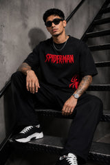 Man wearing a black oversized t-shirt with a red 'SPIDERMAN' puff print graphic and a red spider logo on the side. Model is wearing sunglasses and cargo pants.