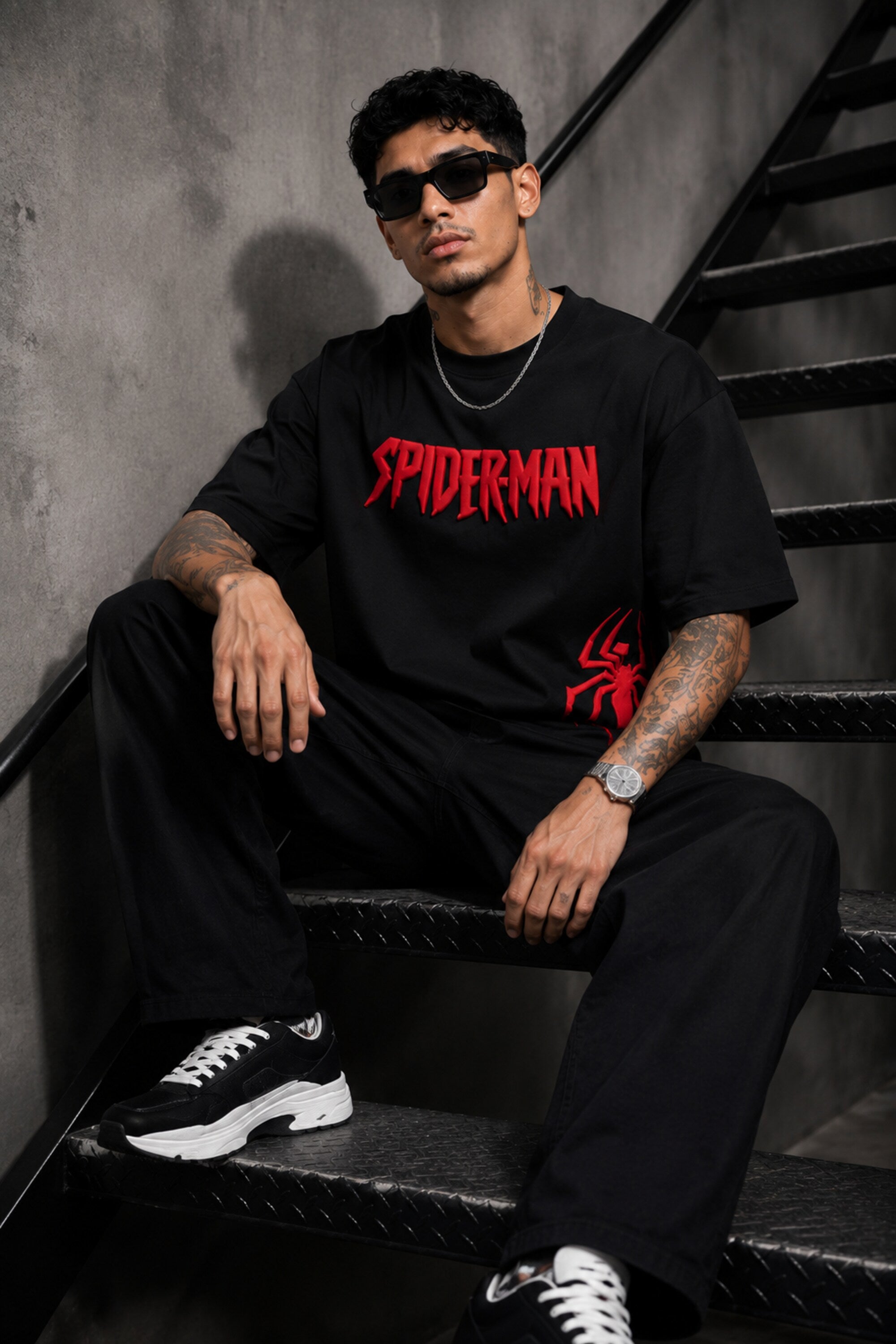 Man wearing a black oversized t-shirt with a red 'SPIDERMAN' puff print graphic and a red spider logo on the side. Model is wearing sunglasses and cargo pants.