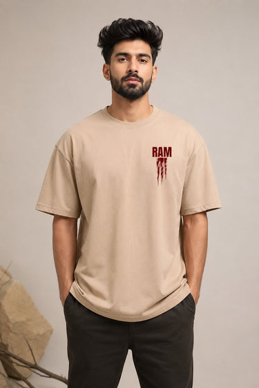 Model wearing a beige RAM oversized t-shirt with a red graphic print on the chest, paired with dark trousers and white sneakers. Showcases streetwear style and premium comfort.