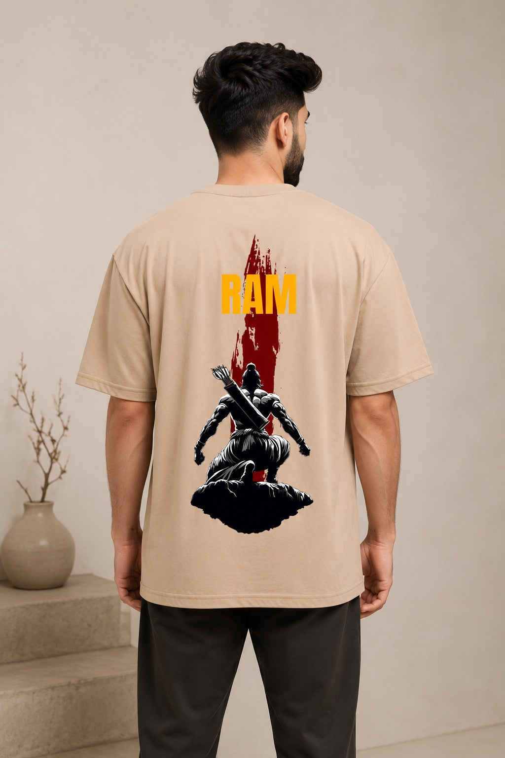Model wearing a beige RAM oversized t-shirt with a red graphic print on the chest, paired with dark trousers and white sneakers. Showcases streetwear style and premium comfort. - beige