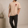 Model wearing a beige RAM oversized t-shirt with a red graphic print on the chest, paired with dark trousers and white sneakers. Showcases streetwear style and premium comfort.