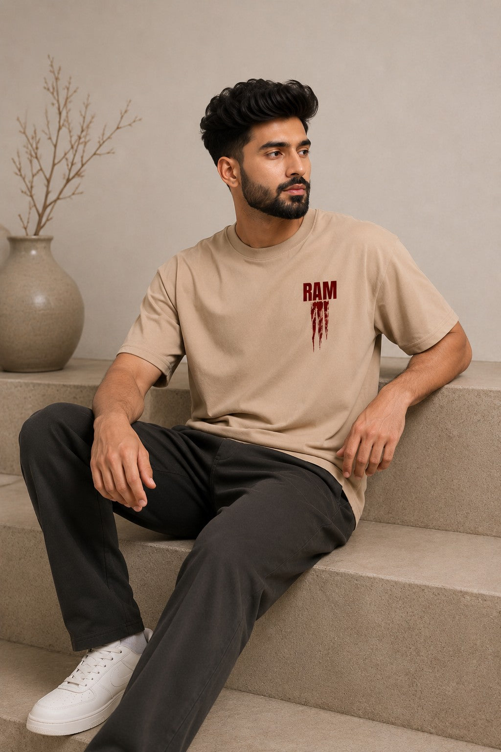 Model wearing a beige RAM oversized t-shirt with a red graphic print on the chest, paired with dark trousers and white sneakers. Showcases streetwear style and premium comfort.