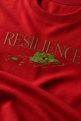 Red oversized t-shirt with "RESILIENCE" embroidered in gold and a green embroidered crocodile graphic. Features a ribbed neckline and premium stitching.