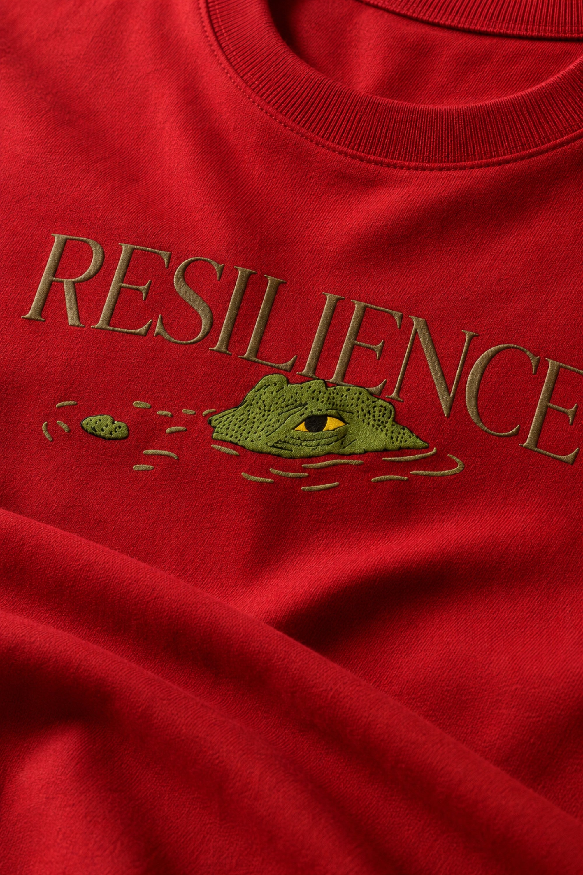 Red oversized t-shirt with "RESILIENCE" embroidered in gold and a green embroidered crocodile graphic. Features a ribbed neckline and premium stitching.