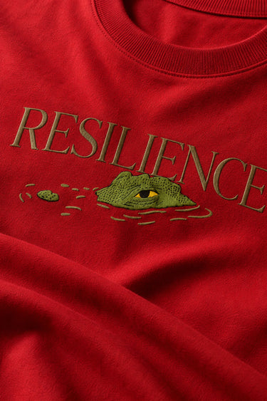 Red oversized t-shirt with "RESILIENCE" embroidered in gold and a green embroidered crocodile graphic. Features a ribbed neckline and premium stitching.