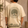 Close-up of a Rooted oversized t-shirt in beige, featuring a dark green circular graphic of a tree of life with tribal figures, and text 'in the rhfym of life'. - beige