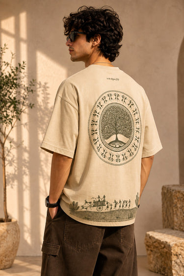 Close-up of a Rooted oversized t-shirt in beige, featuring a dark green circular graphic of a tree of life with tribal figures, and text 'in the rhfym of life'.