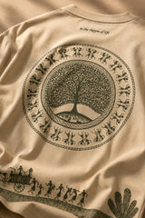 Close-up of a Rooted oversized t-shirt in beige, featuring a dark green circular graphic of a tree of life with tribal figures, and text 'in the rhfym of life'.