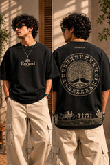 Close-up of a Rooted oversized t-shirt in beige, featuring a dark green circular graphic of a tree of life with tribal figures, and text 'in the rhfym of life'. - black