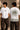 Close-up of a Rooted oversized t-shirt in beige, featuring a dark green circular graphic of a tree of life with tribal figures, and text 'in the rhfym of life'. - white