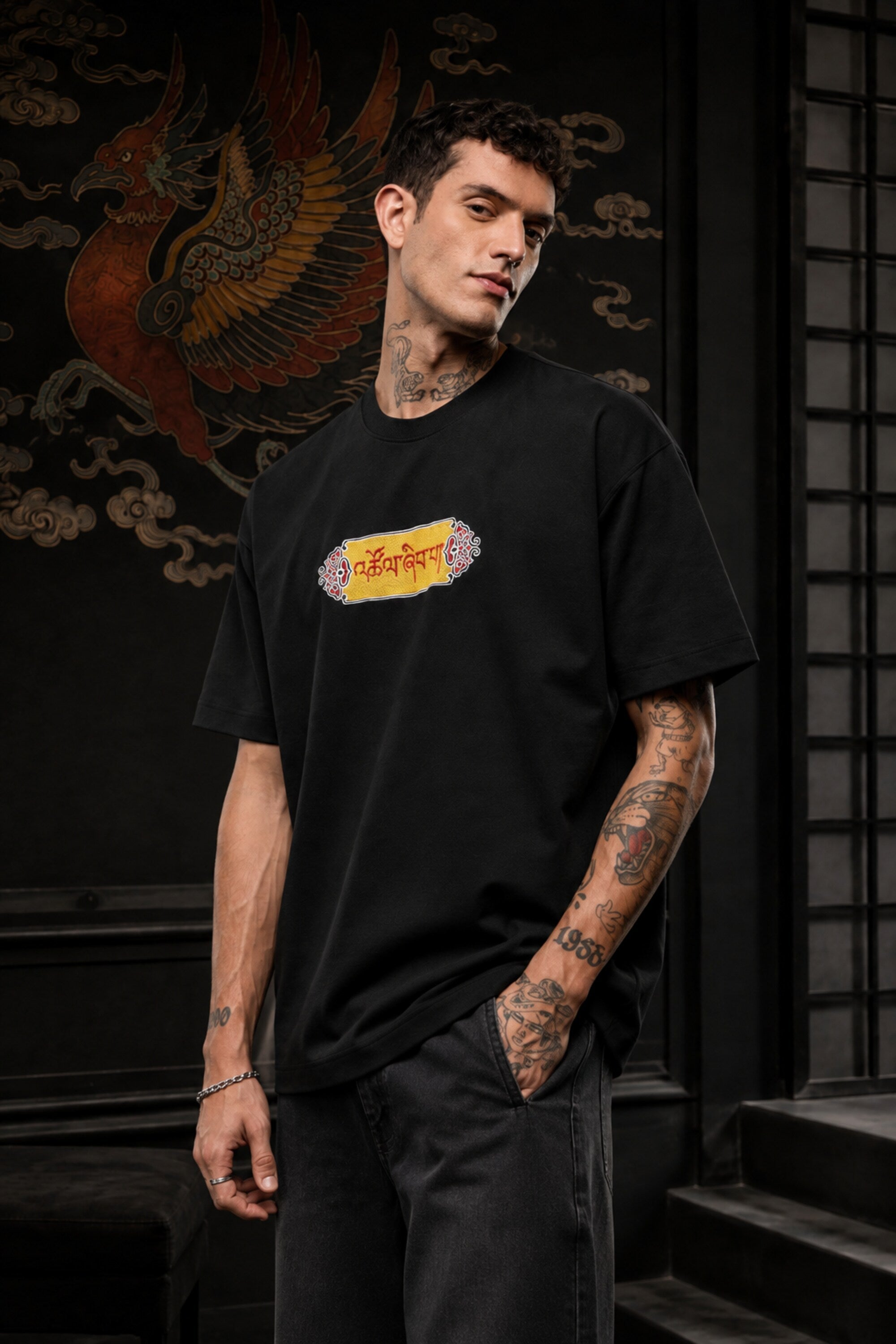Black oversized t-shirt with Tibetan script in red on golden decorative banner, spiritual streetwear India