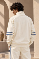 A man wearing a cream-colored athletic tracksuit with navy blue stripes on the sleeves and side, photographed from behind in a sunlit architectural setting.