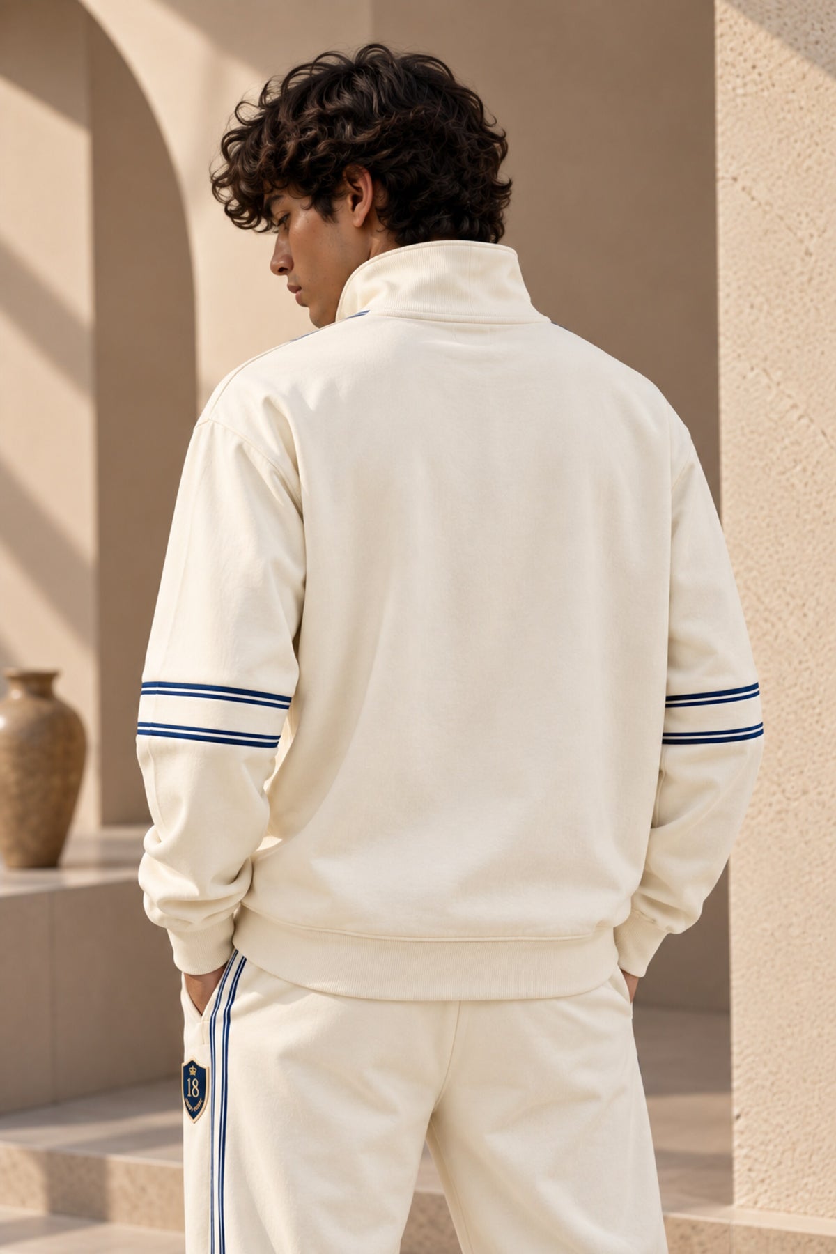 A man wearing a cream-colored athletic tracksuit with navy blue stripes on the sleeves and side, photographed from behind in a sunlit architectural setting.