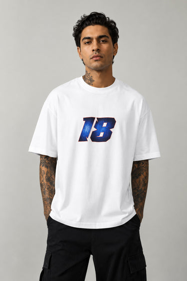 Man wearing a white VIRAT MCG 18 Vs PAK oversized t-shirt with a blue galaxy-themed '18' graphic. He is sitting on steps, showcasing the relaxed fit and streetwear style.