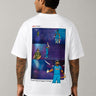 Man wearing a white VIRAT MCG 18 Vs PAK oversized t-shirt with a blue galaxy-themed '18' graphic. He is sitting on steps, showcasing the relaxed fit and streetwear style. - white