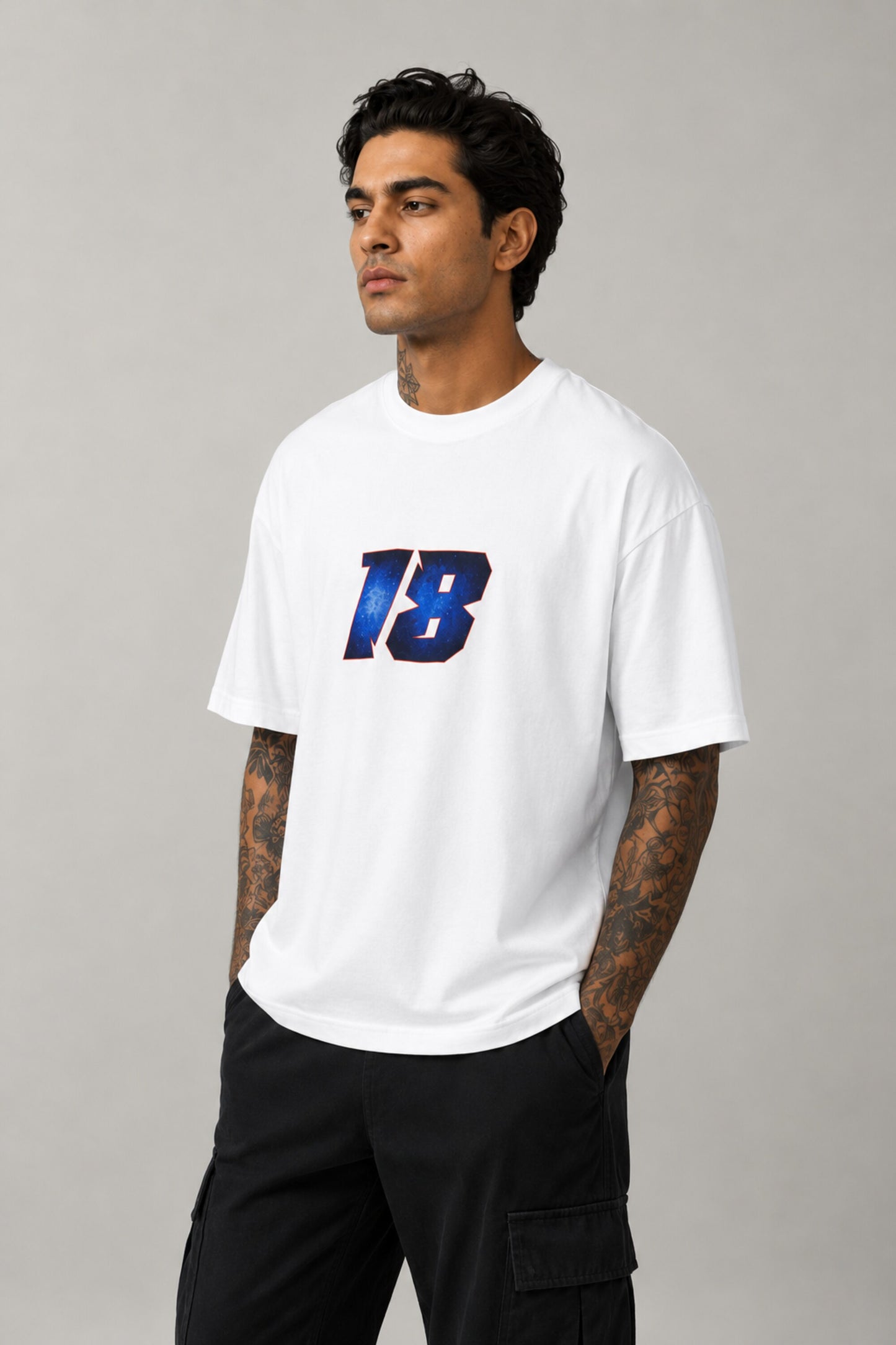 Man wearing a white VIRAT MCG 18 Vs PAK oversized t-shirt with a blue galaxy-themed '18' graphic. He is sitting on steps, showcasing the relaxed fit and streetwear style.