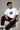Man wearing a white VIRAT MCG 18 Vs PAK oversized t-shirt with a blue galaxy-themed '18' graphic. He is sitting on steps, showcasing the relaxed fit and streetwear style.