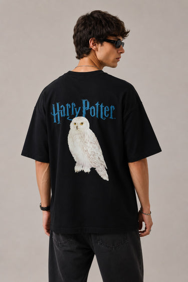 Model wearing a black Harry Potter Gryffindor oversized t-shirt with a blue 'HP' graphic and Hogwarts crest. Features black sunglasses, silver necklace, and dark pants.