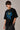 Model wearing a black Harry Potter Gryffindor oversized t-shirt with a blue 'HP' graphic and Hogwarts crest. Features black sunglasses, silver necklace, and dark pants.