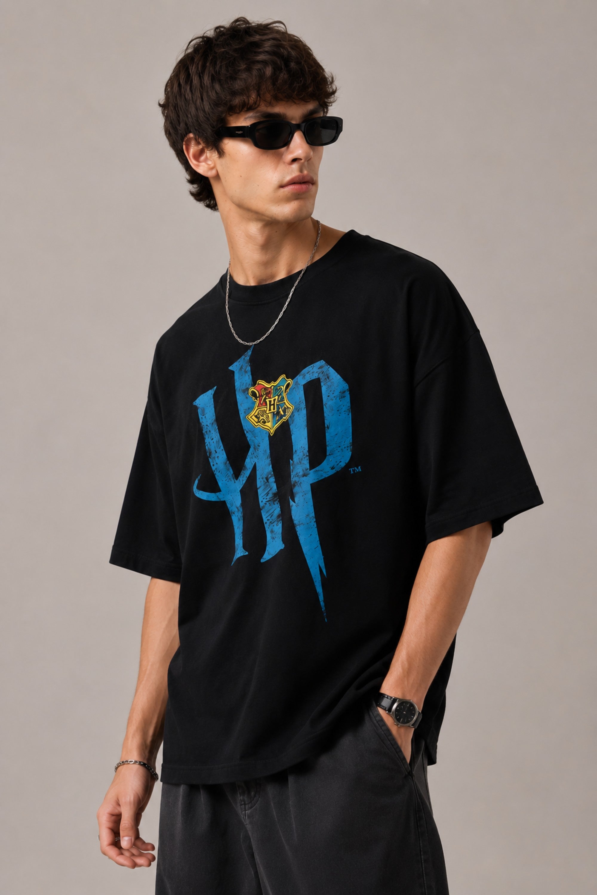 Model wearing a black Harry Potter Gryffindor oversized t-shirt with a blue 'HP' graphic and Hogwarts crest. Features black sunglasses, silver necklace, and dark pants.