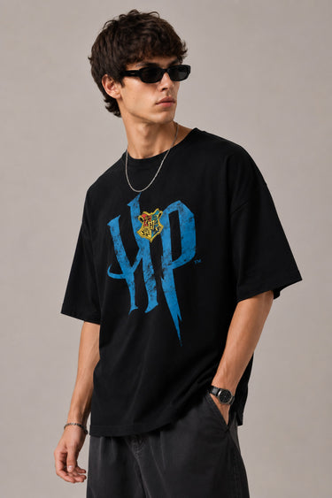 Model wearing a black Harry Potter Gryffindor oversized t-shirt with a blue 'HP' graphic and Hogwarts crest. Features black sunglasses, silver necklace, and dark pants.