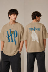 Model wearing a black Harry Potter Gryffindor oversized t-shirt with a blue 'HP' graphic and Hogwarts crest. Features black sunglasses, silver necklace, and dark pants. - beige