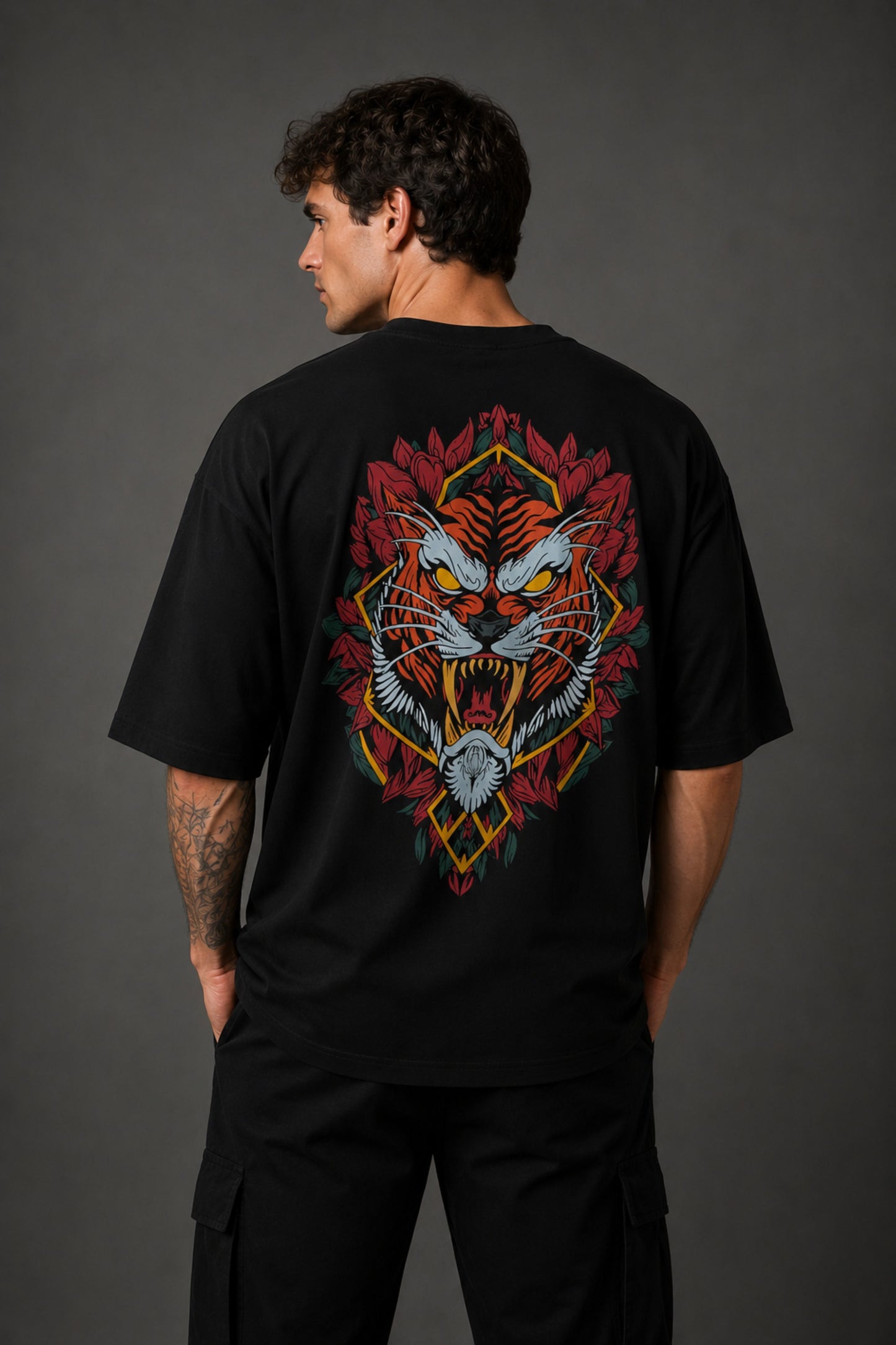 Man wearing a black UNSTOPPABLE DESIGN oversized t-shirt with a vibrant, detailed graphic of a roaring tiger surrounded by red flowers and a geometric frame on the back.