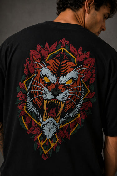 Man wearing a black UNSTOPPABLE DESIGN oversized t-shirt with a vibrant, detailed graphic of a roaring tiger surrounded by red flowers and a geometric frame on the back.